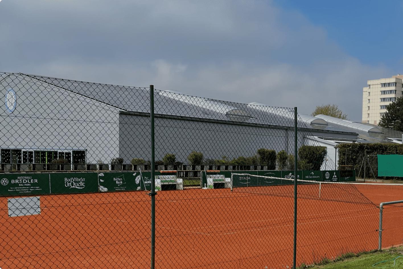 Panoramablick auf einen Tennisplatz mit rotem Sand, umgeben von einem Zaun und einem modernen Gebäude im Hintergrund.